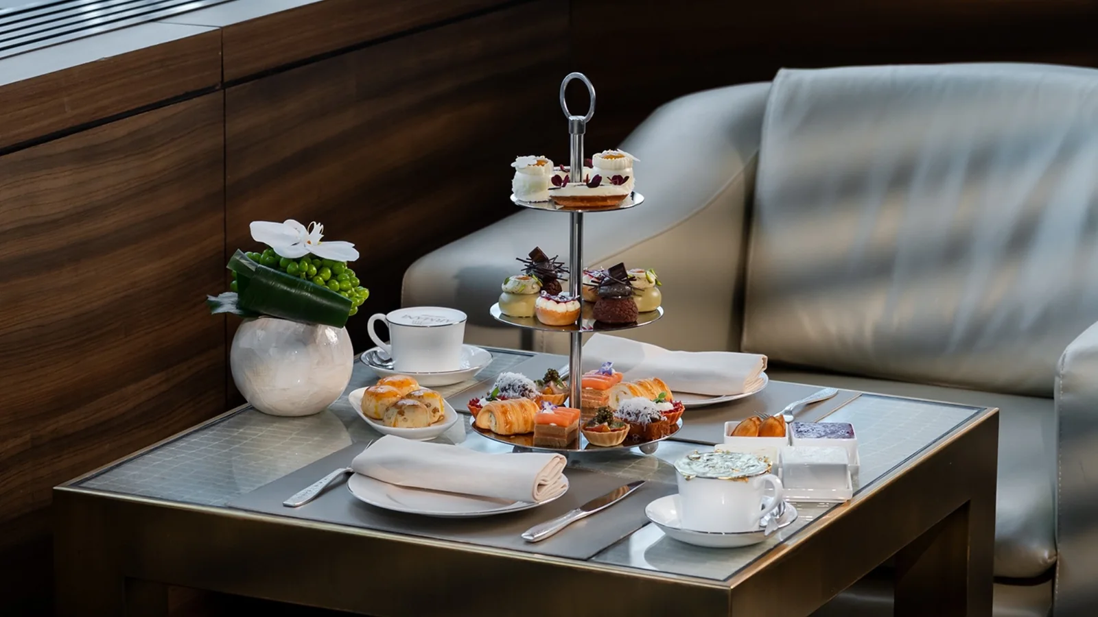 Afternoon tea on a low table beside a cream armchair: a tiered stand of pastries and savouries with teacups, scones and a floral arrangement