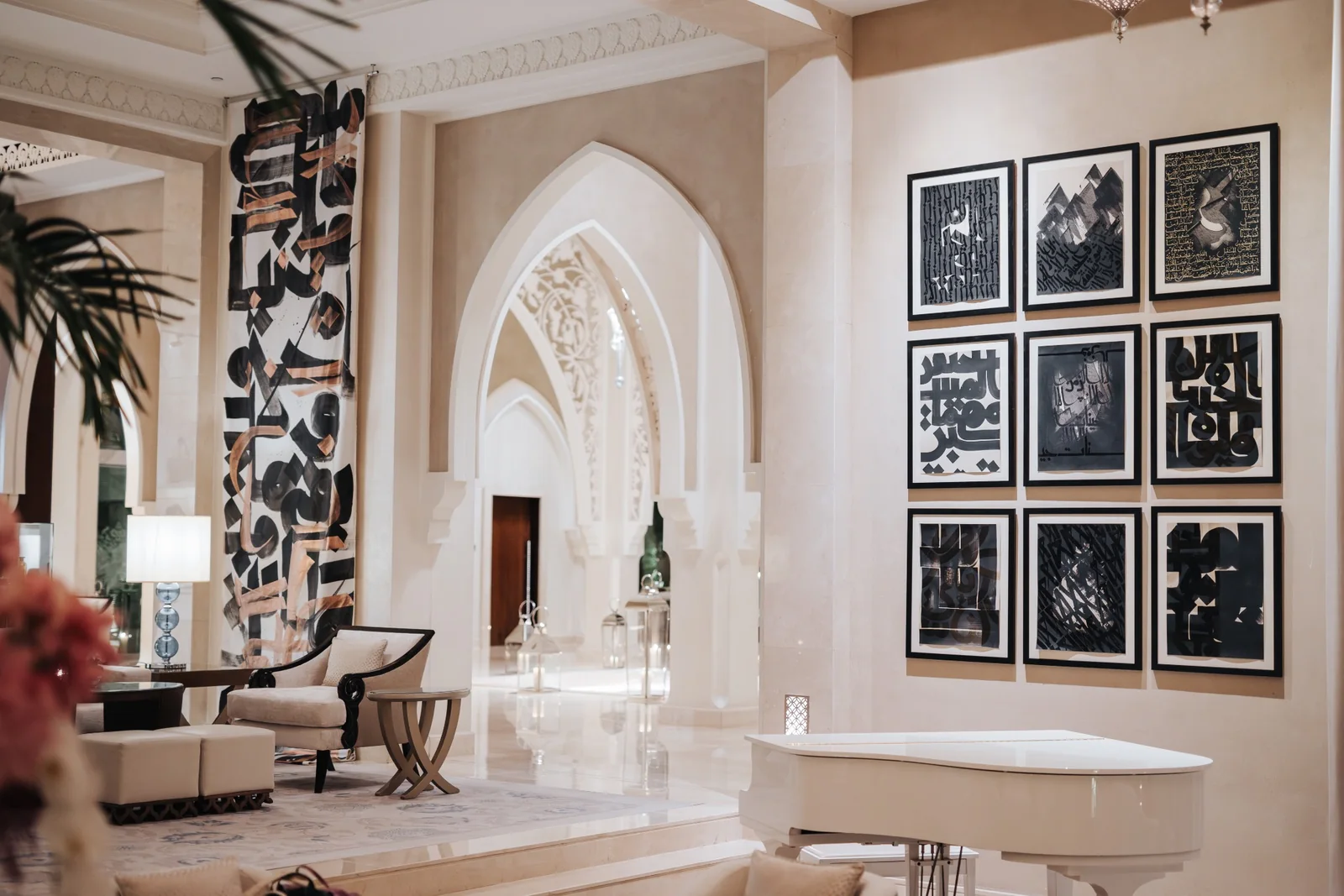 An interior at One&Only The Palm with pointed arches, a tall calligraphy hanging, a grid of framed artworks and a white grand piano