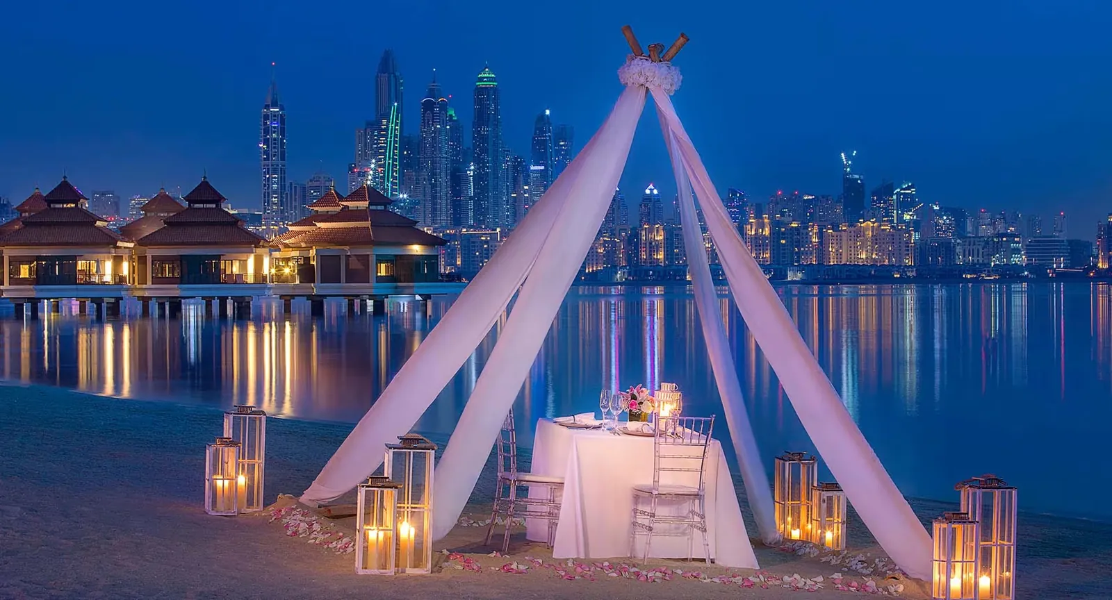 A candlelit table for two under a draped canopy on the beach at night, with over-water villas and the lit Dubai Marina skyline across the water