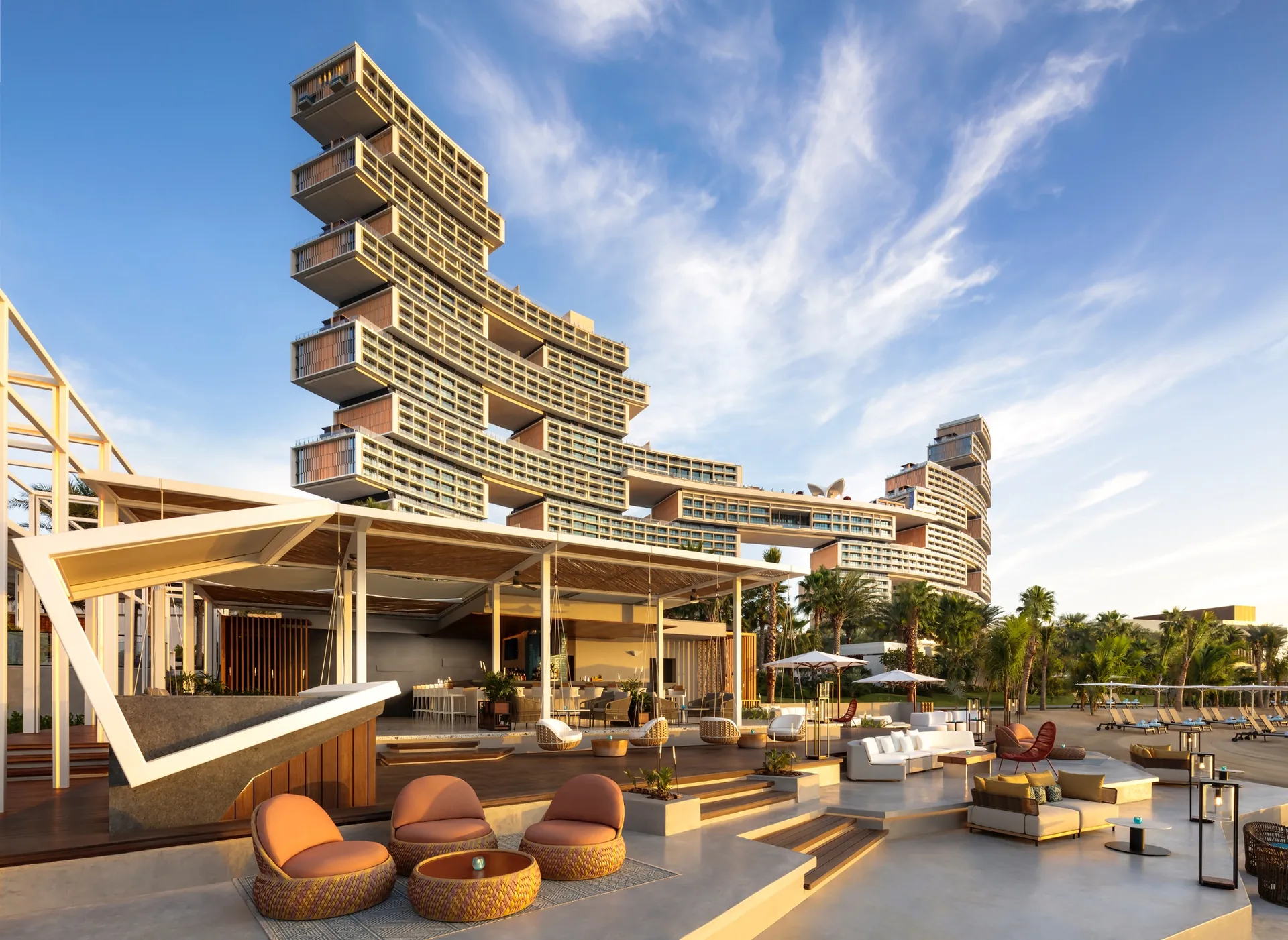 Atlantis The Royal on Palm Jumeirah at golden hour, its stacked offset blocks rising above a shaded beachfront lounge terrace with palms and daybeds