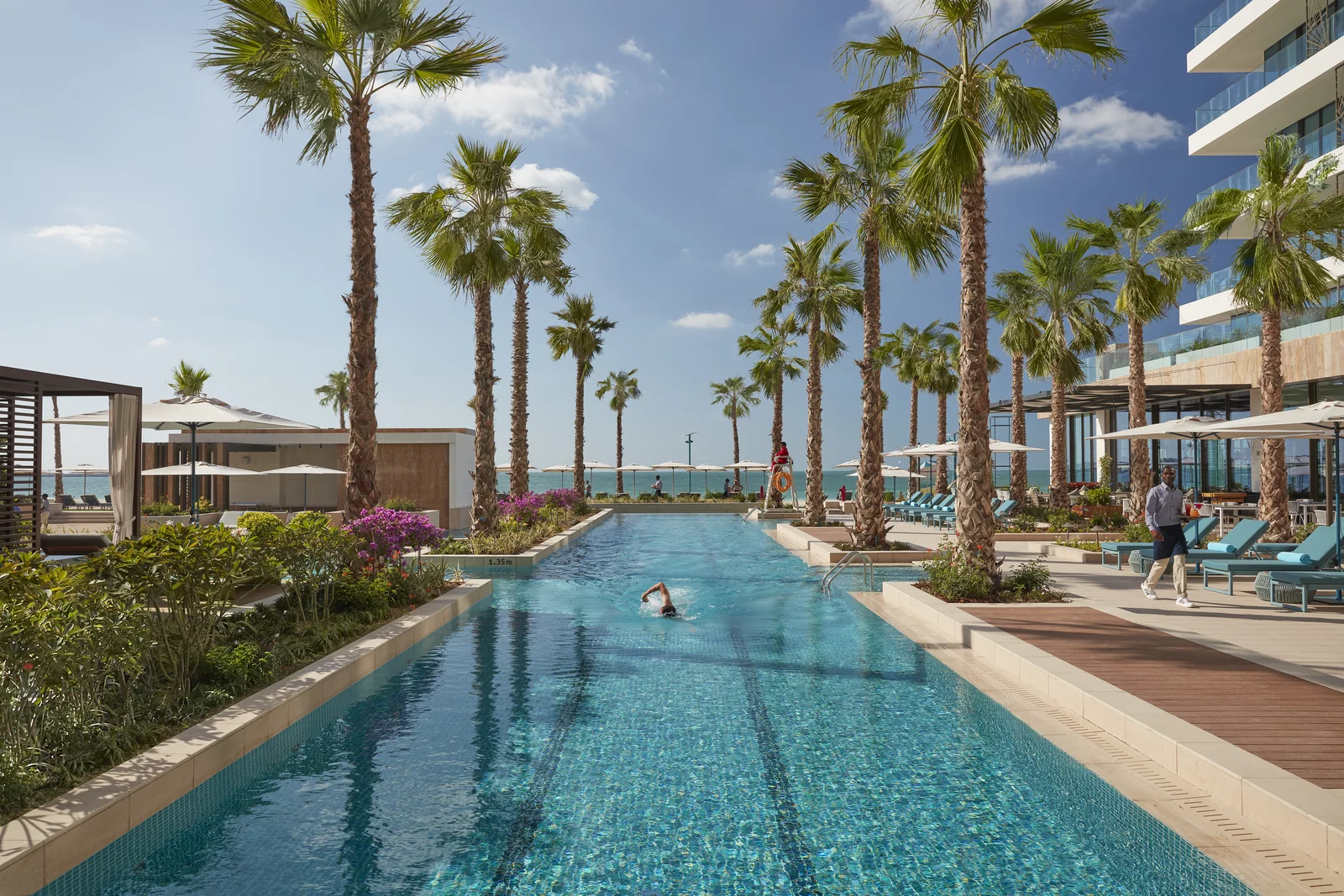 A long lap pool lined with tall palms, loungers and parasols, leading toward the sea