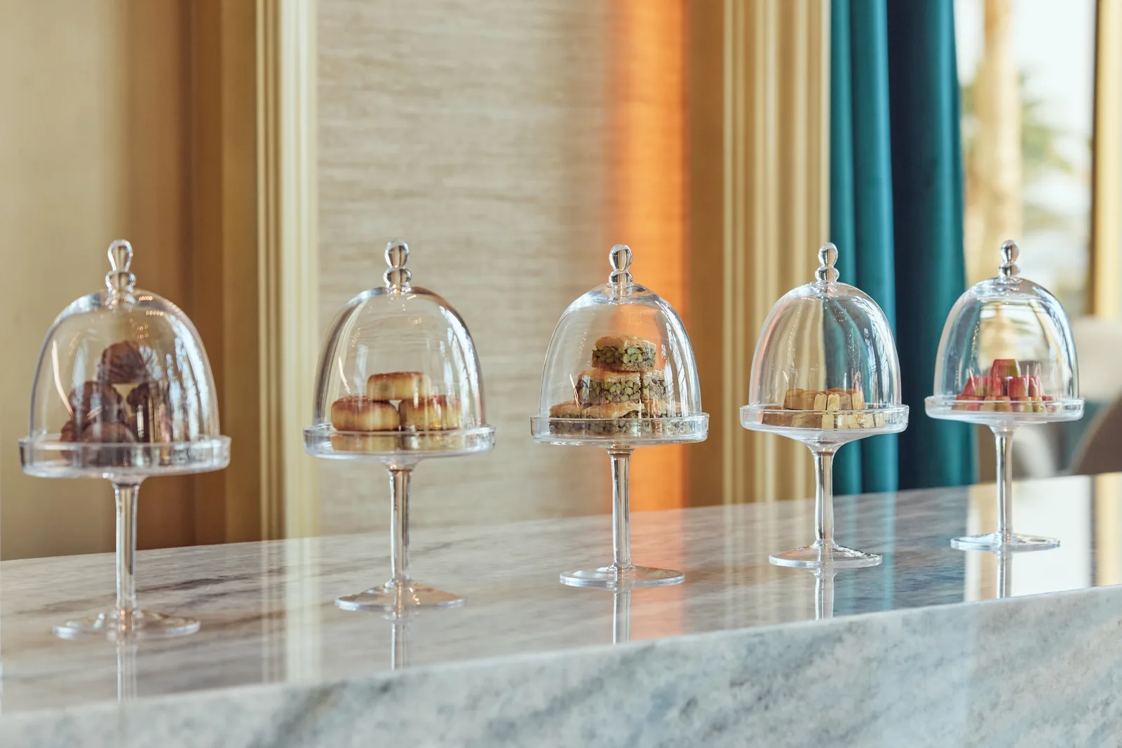 Five glass cloches on stands along a marble counter, each holding pastries and small bites