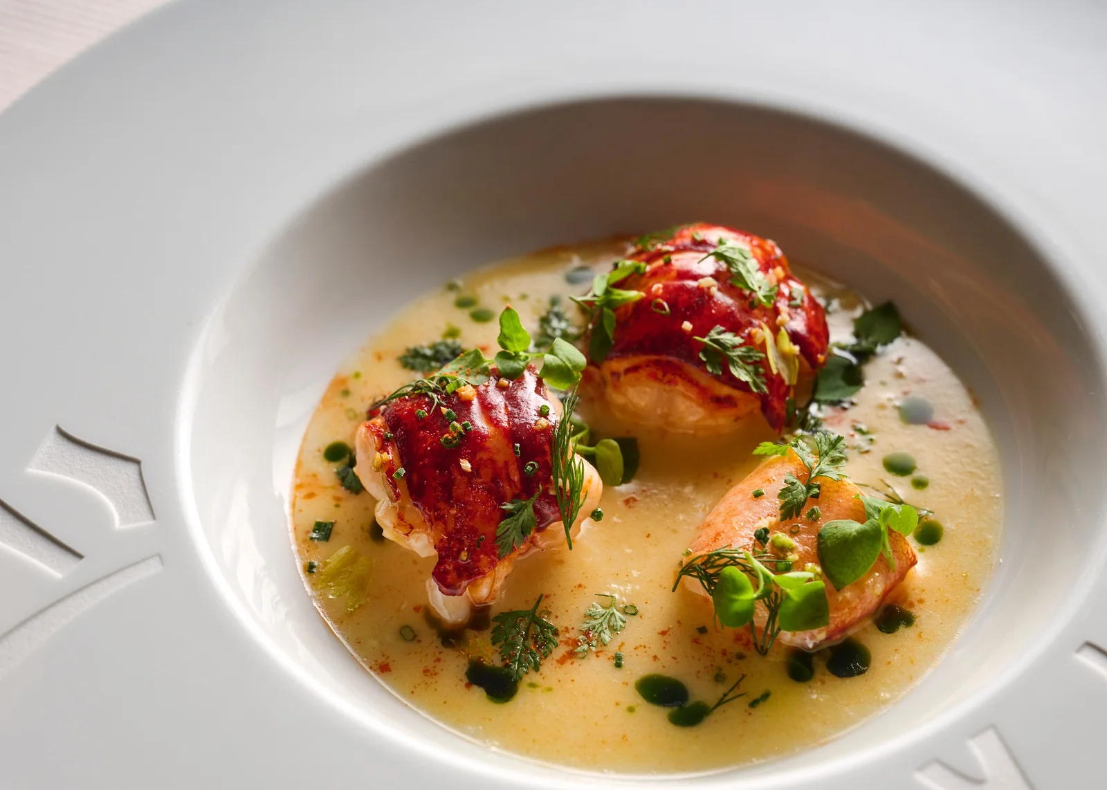 A white bowl holding three pieces of lobster in a pale sauce, finished with herbs and green oil