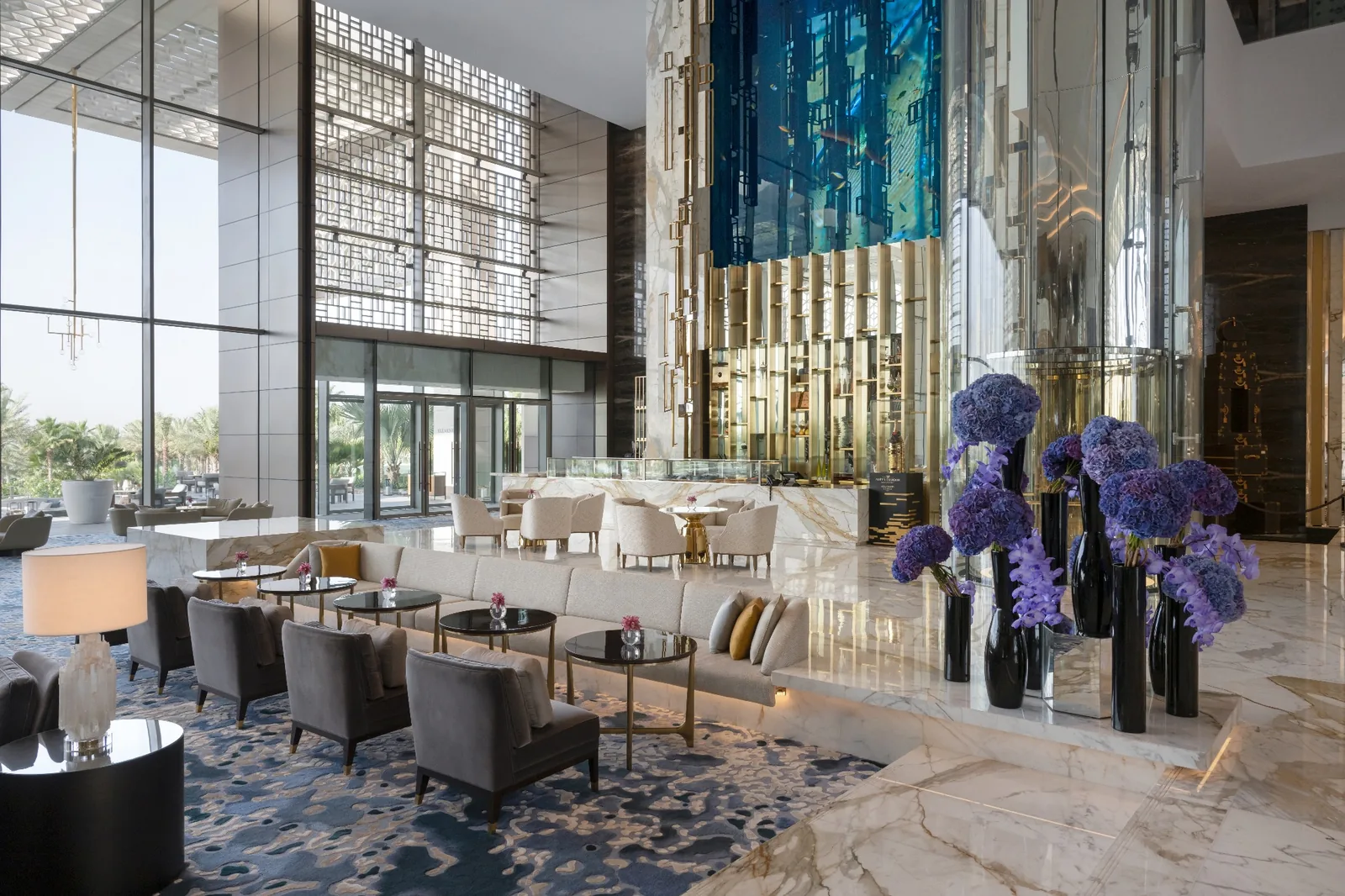 The Royal Tea Room lounge at Atlantis The Royal, a double-height marble interior with brass detailing, blue patterned carpet and tall vases of purple hydrangeas