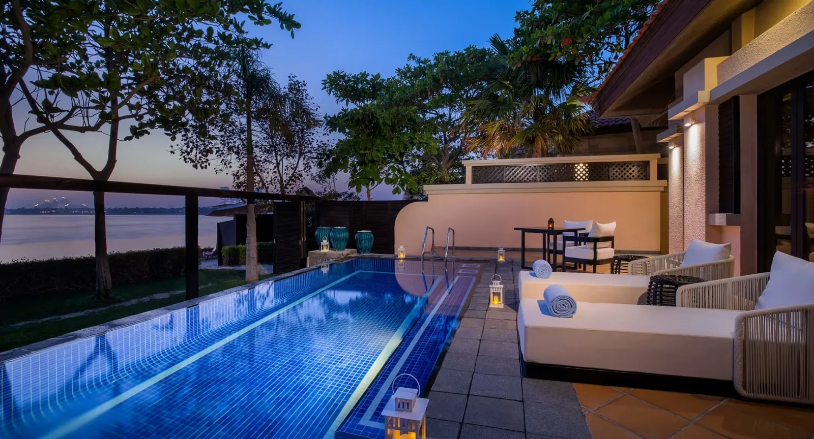 A villa terrace at dusk with a long lit private pool, daybeds, lanterns and trees framing the water beyond
