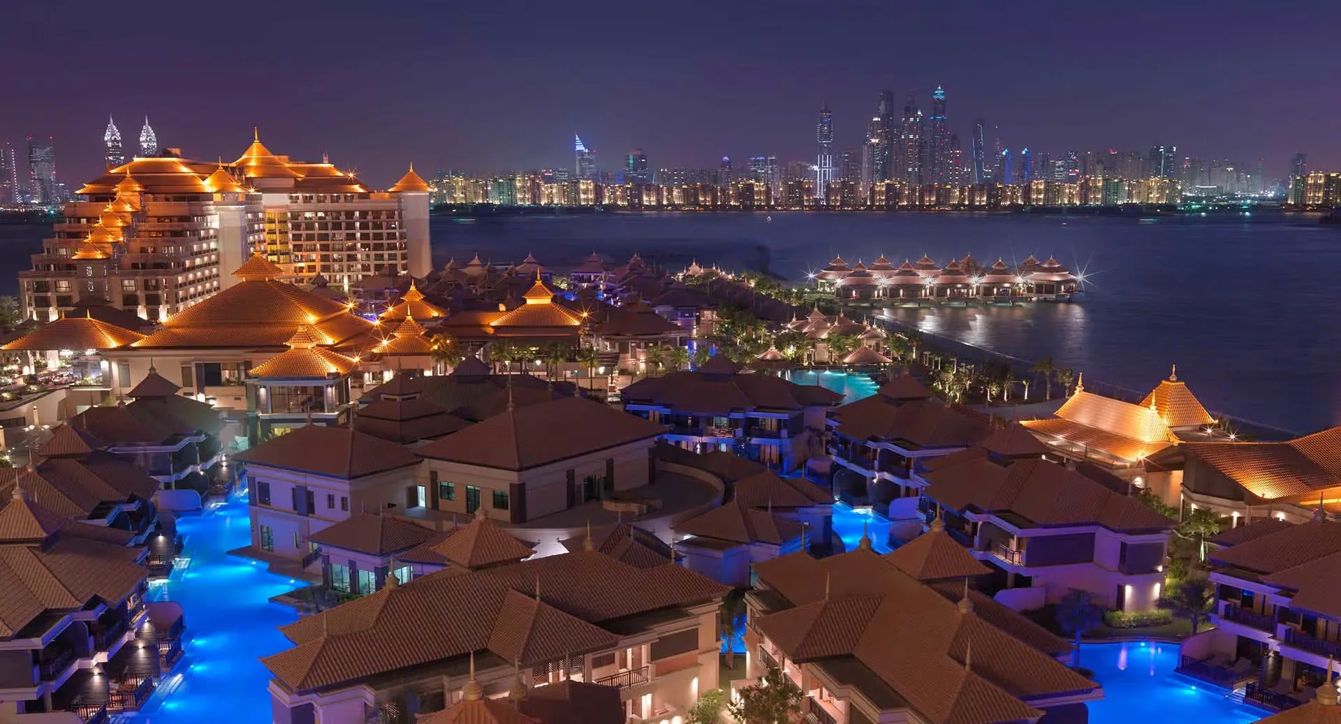 Anantara The Palm Dubai Resort at night, seen from above: tiled pavilion roofs and lit swimming lagoons with over-water villas and the Dubai Marina skyline beyond