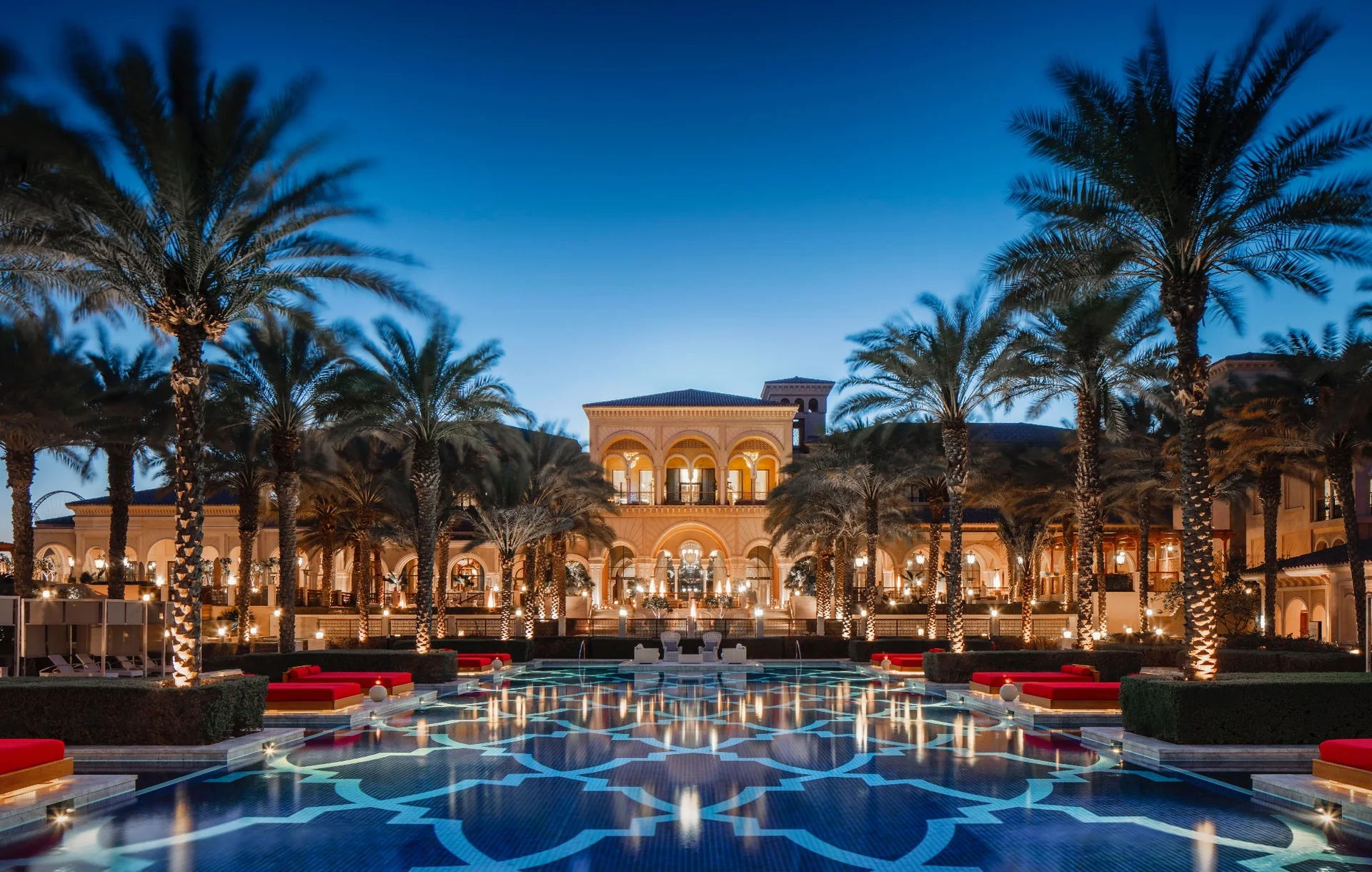 One&Only The Palm at dusk: a patterned mosaic reflecting pool lined with lit palms and red daybeds leading to the arcaded main building